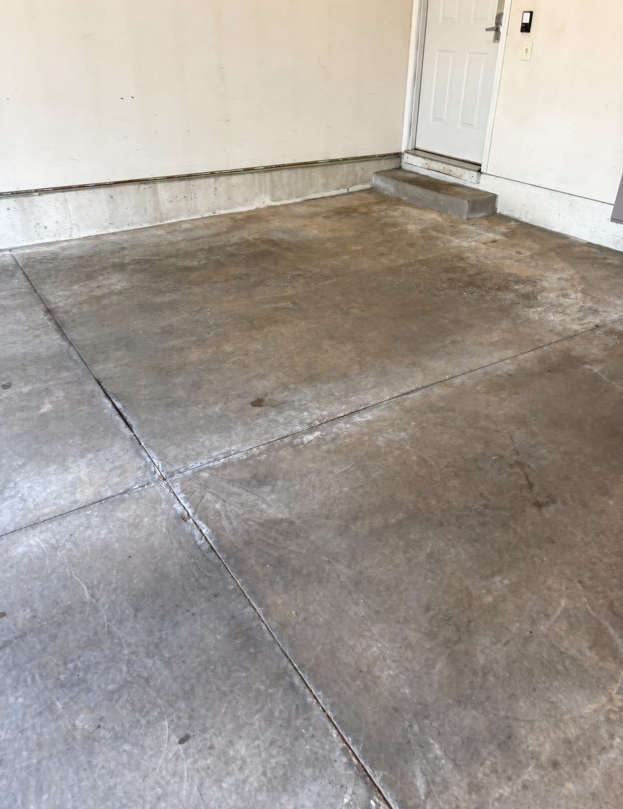 Bare concrete garage floor before coating — grey, stained and worn, with open control joints running across the slab and an uncoated step.